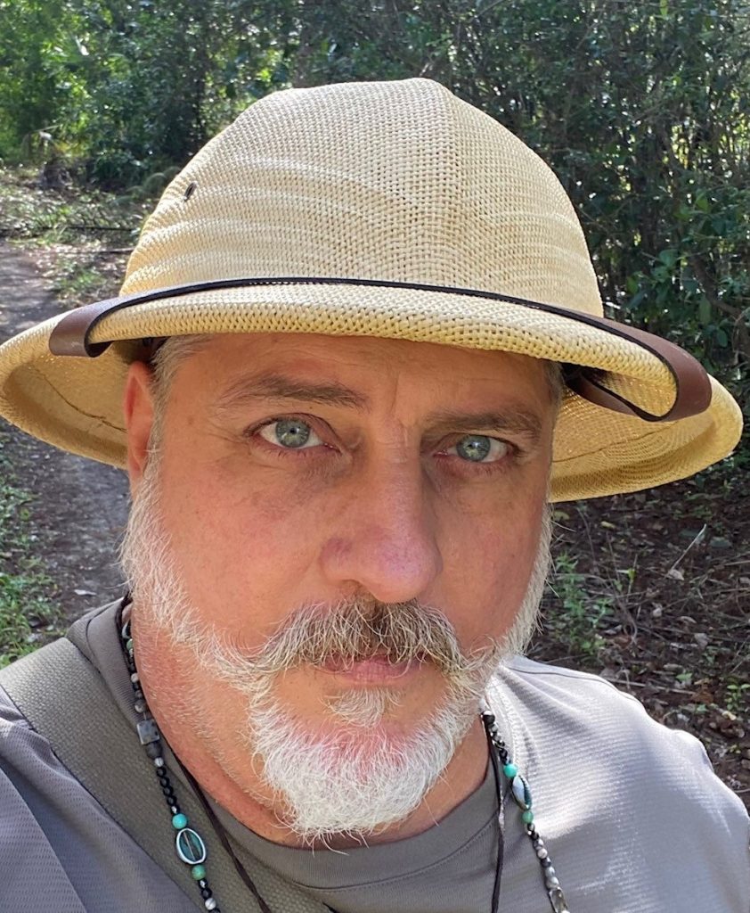 Headshot of a man with a white beard, grey shirt, and tan pith helmet.