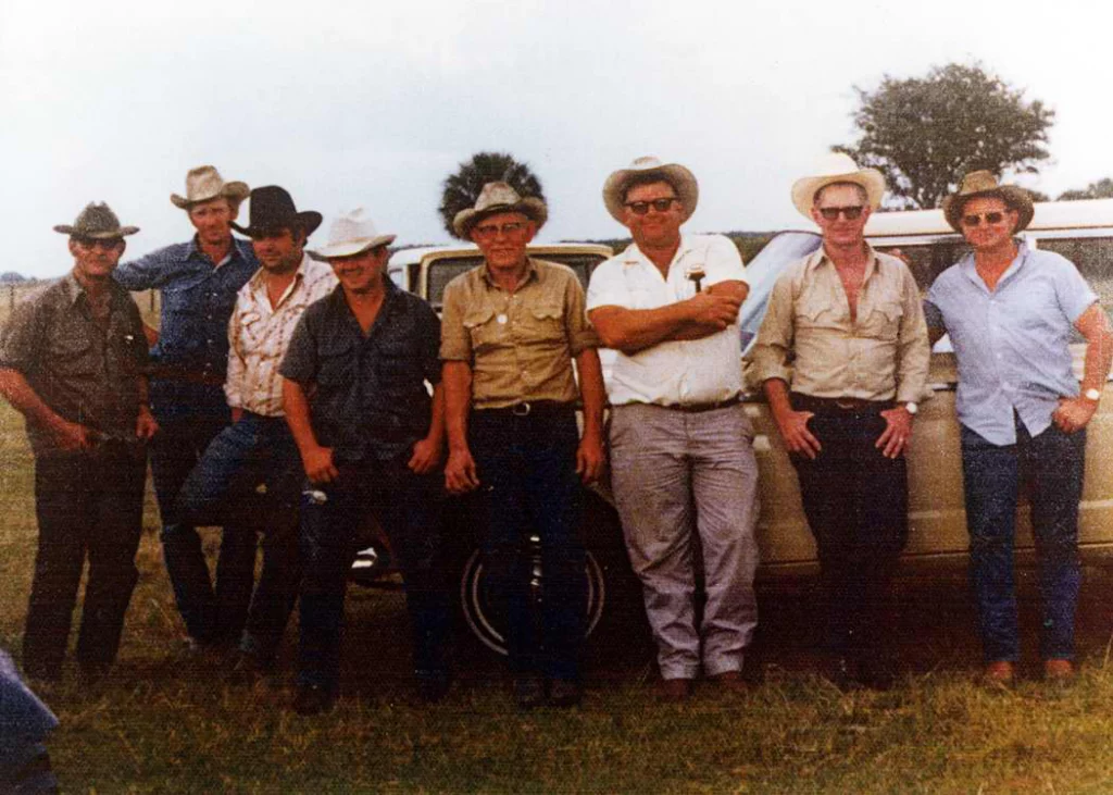 Eight cowboys leaning against a car.