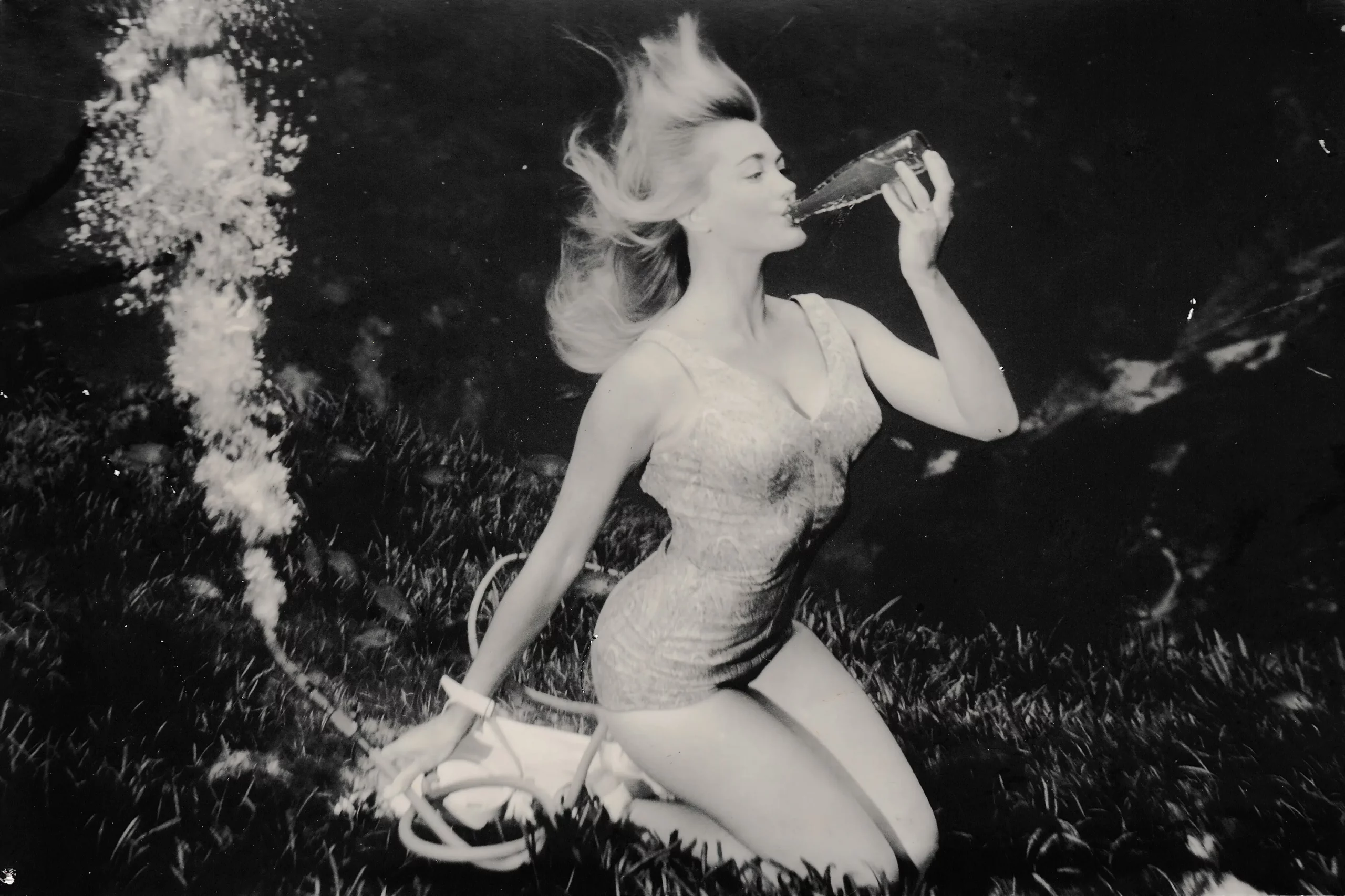 Black and white, a woman in a bathing suit blows into a bottle underwater with an air tube bubbling behind her