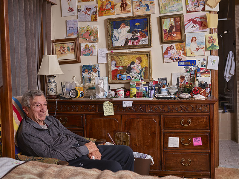 A man with tan skin and medium grey hair seated in front of a cluttered dresser and impressionist art