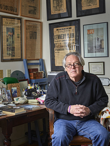 A man with light skin, grey hair, and glasses sitting in front of a cluttered desk in front of framed news articles